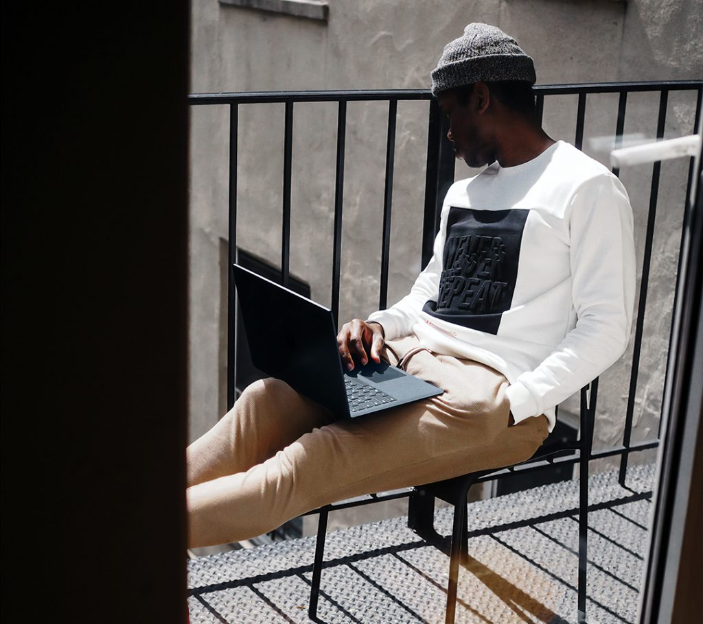 En kille som sitter på en balkong med en laptop i knät. Tittar ut över balkongräcket.
