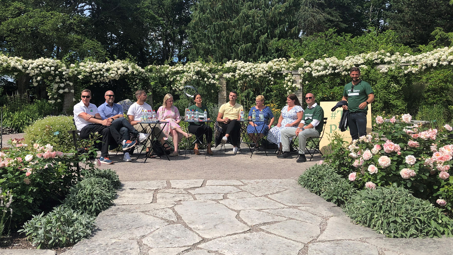 Åtta personer som sitter och en som står upp i en trädgård, omgiven av buskar och blommor. En av personerna pratar och artikulerar med händerna.
