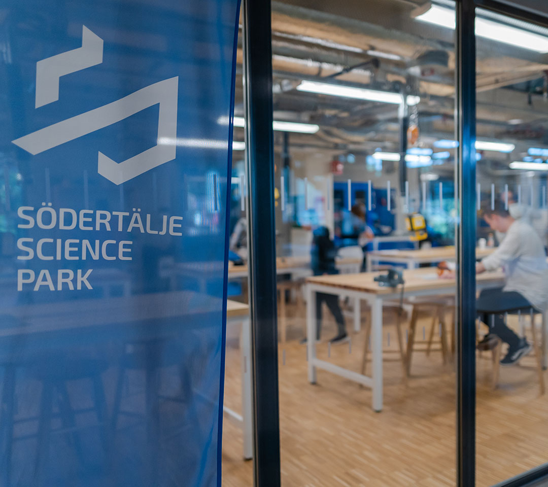 Glasvägg med en vepa som det står Södertälje Sciencepark på. På andra sidan glasväggen är ett produktionsrum, där en person sitter vid ett bord och arbetar.