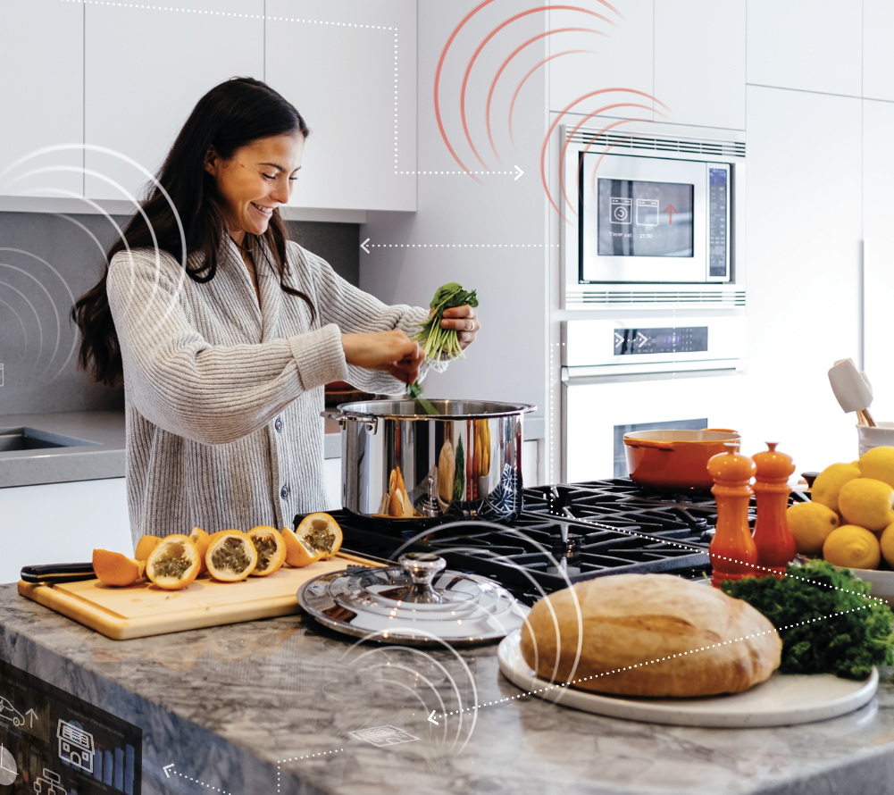 Inomhus i ett kök. En kvinna står och lagar mat på en köksö. På bilden finns illustrerade ikoner som visar signaler och digitalisering.