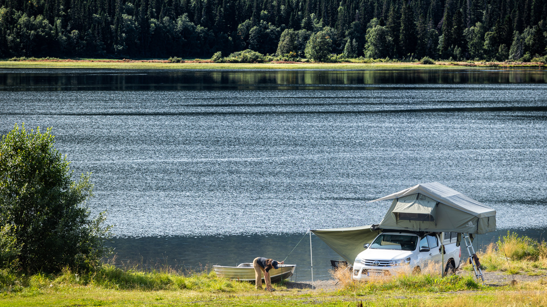 Utomhus. Sommar. En bil med ett taktält som står parkerad vid en sjö. En tjej står bredvid och håller på att sätta upp tältet.